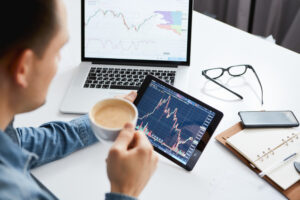 Foto de homem segurando tablet com gráfico de ações e tomando café, com computador, óculos e papéis sobre uma mesa ao fundo.