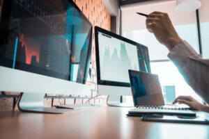 Foto de computador e dois monitores com gráficos de ações