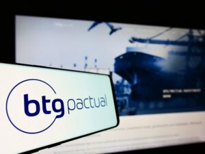 Foto de celular com logo do BTG Pactual e tela de computador ao fundo