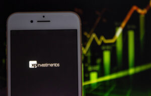 Foto de celular com logo da XP na tela, gráfico de ações ao fundo
