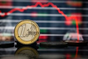 moeda de um euro no primeiro plano com gráfico de preços mostrando queda ao fundo
