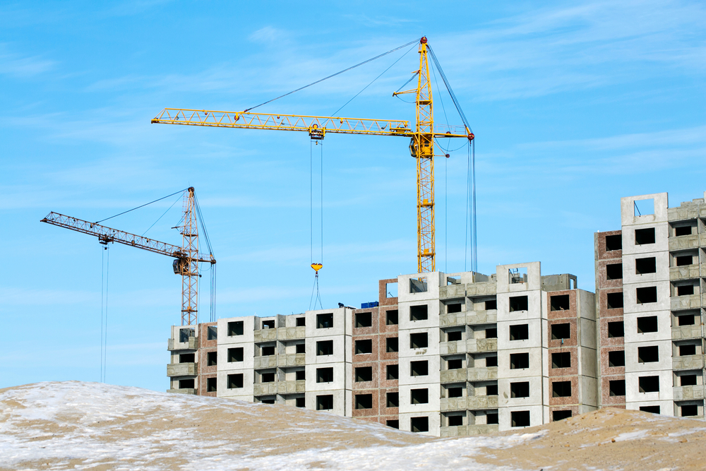 Dois prédios em construção ao fundo da imagem, junto com dois guindastes. À frente, um terreno ainda descampado.