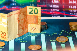 nota de real em primeiro plano e moedas de real com gráfico de colunas e cotações financeiras no segundo plano