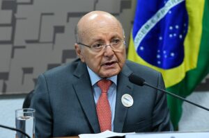 Senhor calvo, de óculos e terno escuro sentado em frente um microfone
