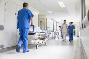 Corredor de hospital com um funcionário de costas, vestido de azul, empurrando uma maca e, na direção contrária, outros dois funcionários caminham, um de branco, outro azul.