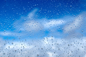 Imagem de céu azul com nuvens atrás de vidro com pingos de chuva.